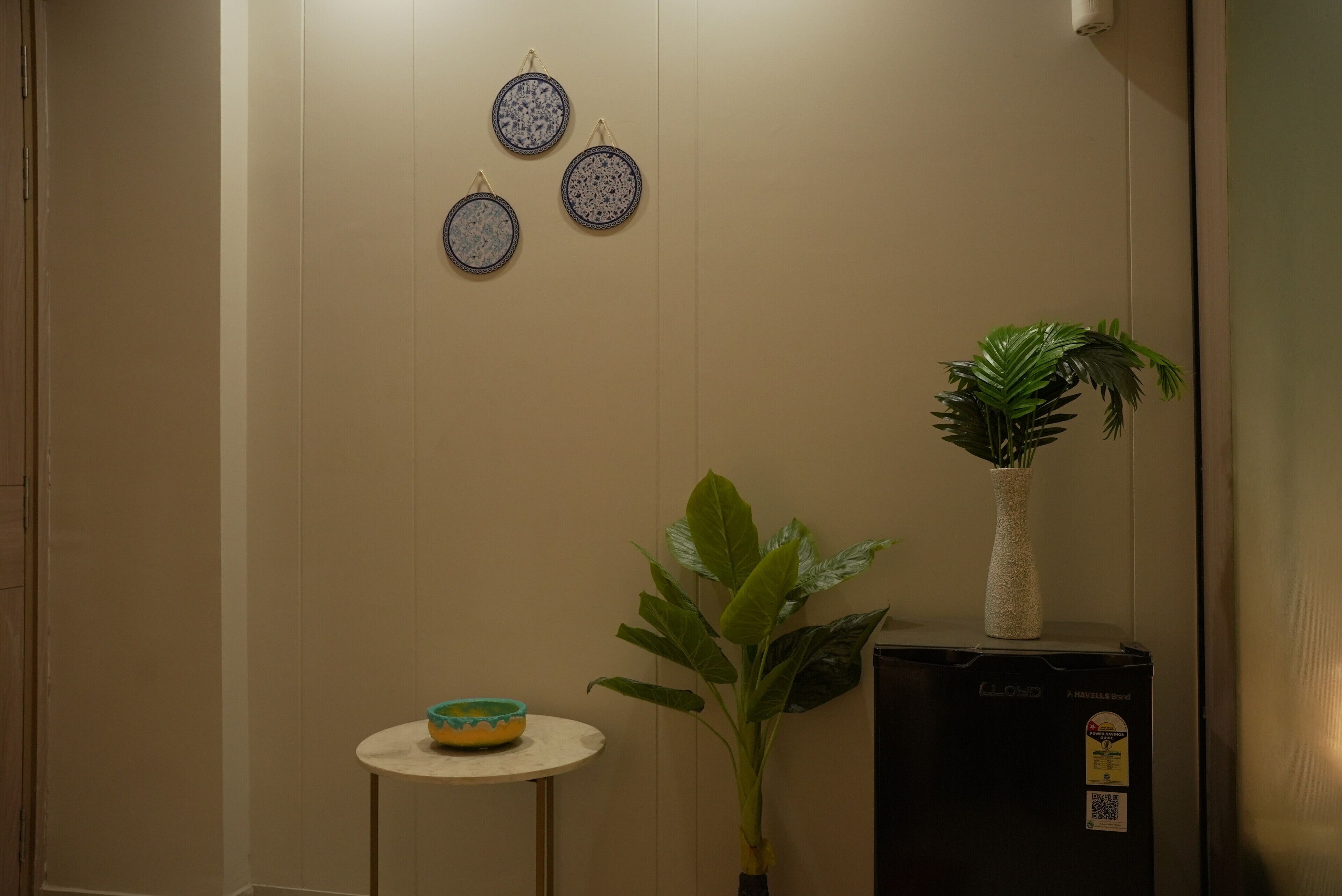 A room with a beige wall, a small round table with a bowl, a potted plant, and a black appliance with a label.