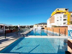 Una piscina amb una superfície d'aigua blava i cristal·lina, envoltada per una tanca i adossada a un edifici amb façana groga i blanca.