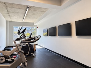 A gym with exercise equipment, a large window, and three mounted screens on the wall. - Indoor Spa | Gym | 9th Floor | Urban Oasis (Québec)