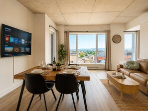 Una sala de estar moderna con comedor, un televisor de pantalla plana mostrando servicios de streaming y vistas de la ciudad a través de ventanas amplias.
