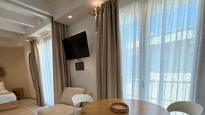 A modern hotel room with a round dining table, two white chairs, a sofa, a flat-screen TV mounted on the wall, and large windows with curtains.