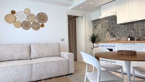 A modern living room with a sofa, a dining table with chairs, and a kitchen area with cabinets and a patterned backsplash.
