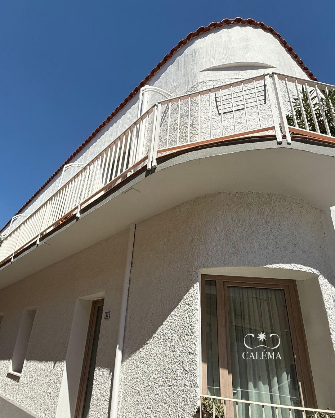 Um edifício com fachada branca, uma varanda com grades brancas e uma janela com cortina exibindo a palavra 'CALÉMA'.