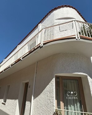 Un edificio con fachada blanca, un balcón con barandilla blanca y una ventana con cortina que muestra la palabra 'CALÉMA'.