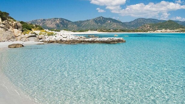 A beach with clear turquoise water, a rocky shoreline, and mountains in the background.
