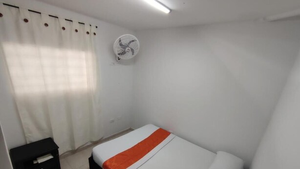 A small, single-bed room with a white bedspread, a black nightstand, a white wall fan, and a window with white curtains. - Gaia Rodadero (Santa Marta)