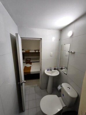 Pieni kylpyhuone, jossa on wc, pesuallas ja peili. Taustalla näkyy pieni sänky.