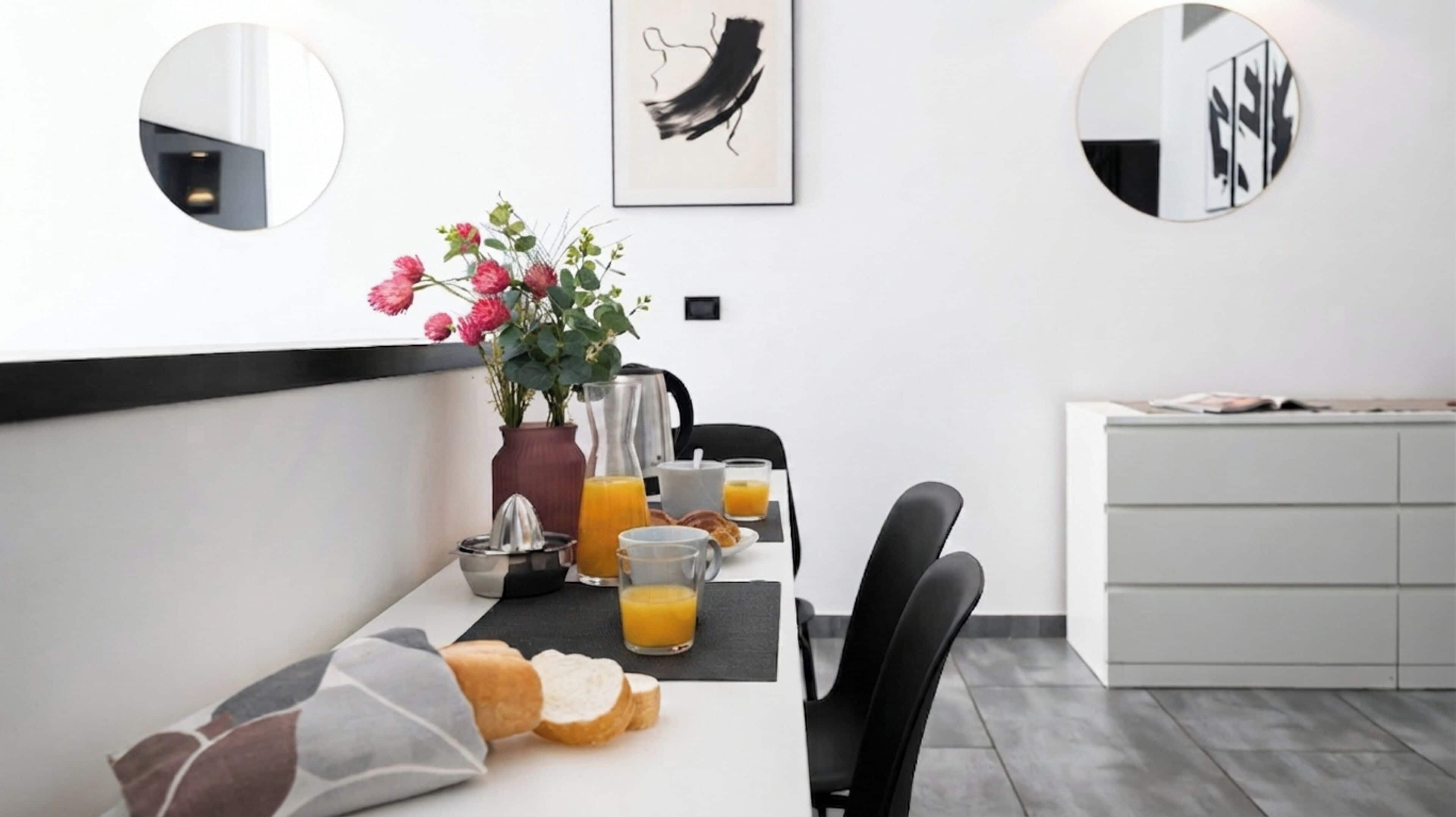 A modern dining area with a long table, chairs, a vase with flowers, a coffee maker, and breakfast items.