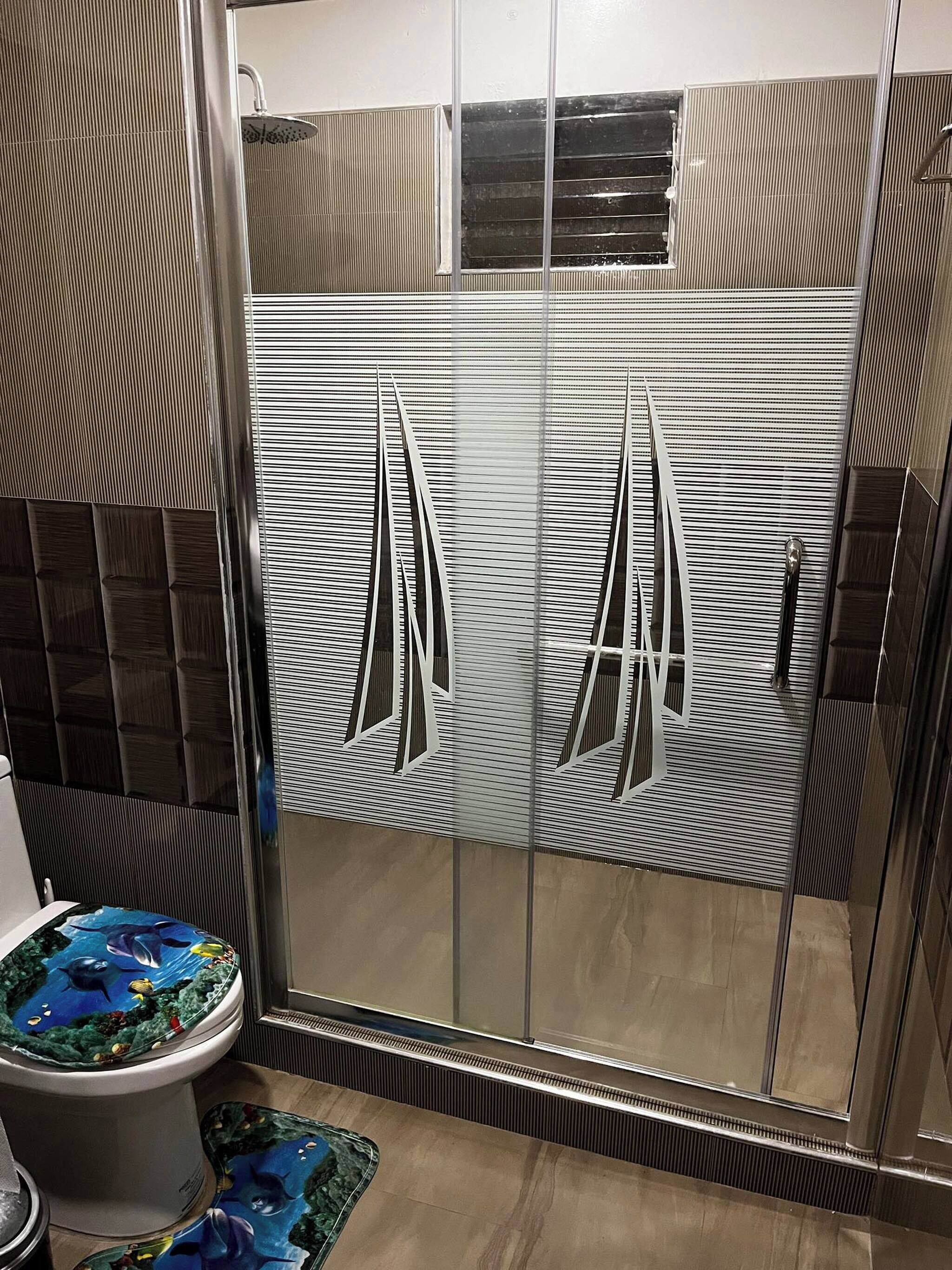 A bathroom with a glass shower enclosure, a toilet with a decorative cover, and a floor with a patterned mat.