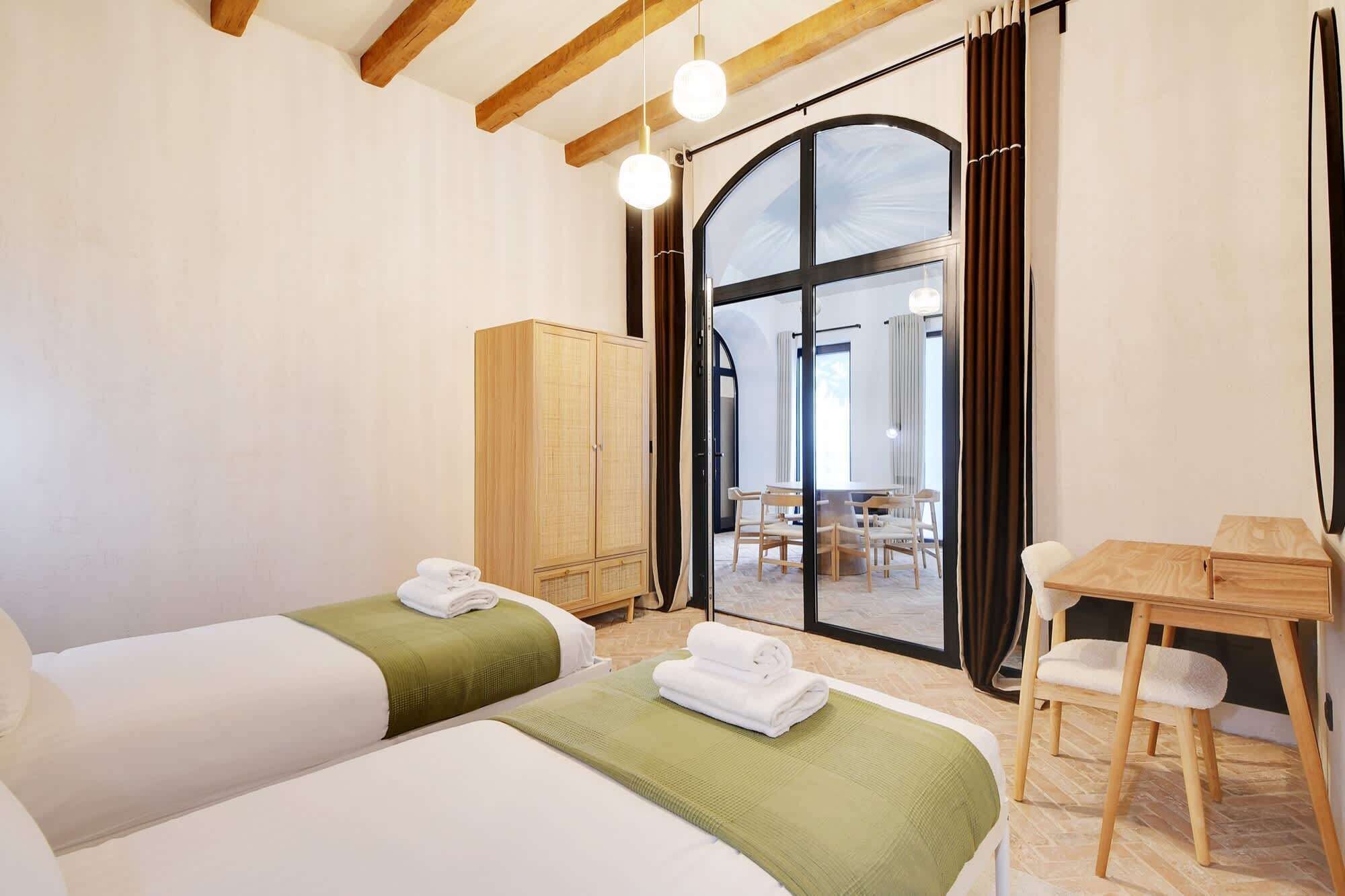 A hotel room with two beds, a wooden wardrobe, a dining table with chairs, and a large arched window.