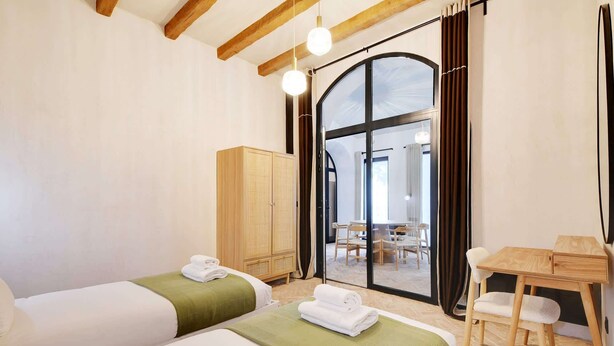A hotel room with two beds, a wooden wardrobe, a dining table with chairs, and a large arched window. - A Authentic 3-BR/2Ba in Opera - Grands Boulevards (Paris)