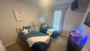 Un dormitorio con dos camas, una mesita de noche, una lámpara, una ventana con cortinas y un televisor montado en la pared.