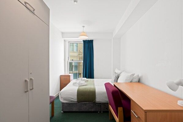 A compact hotel room with a bed, a desk, a chair, a wardrobe, and a window with curtains. - Kings Residence 2 | By My Getaways (Brighton)