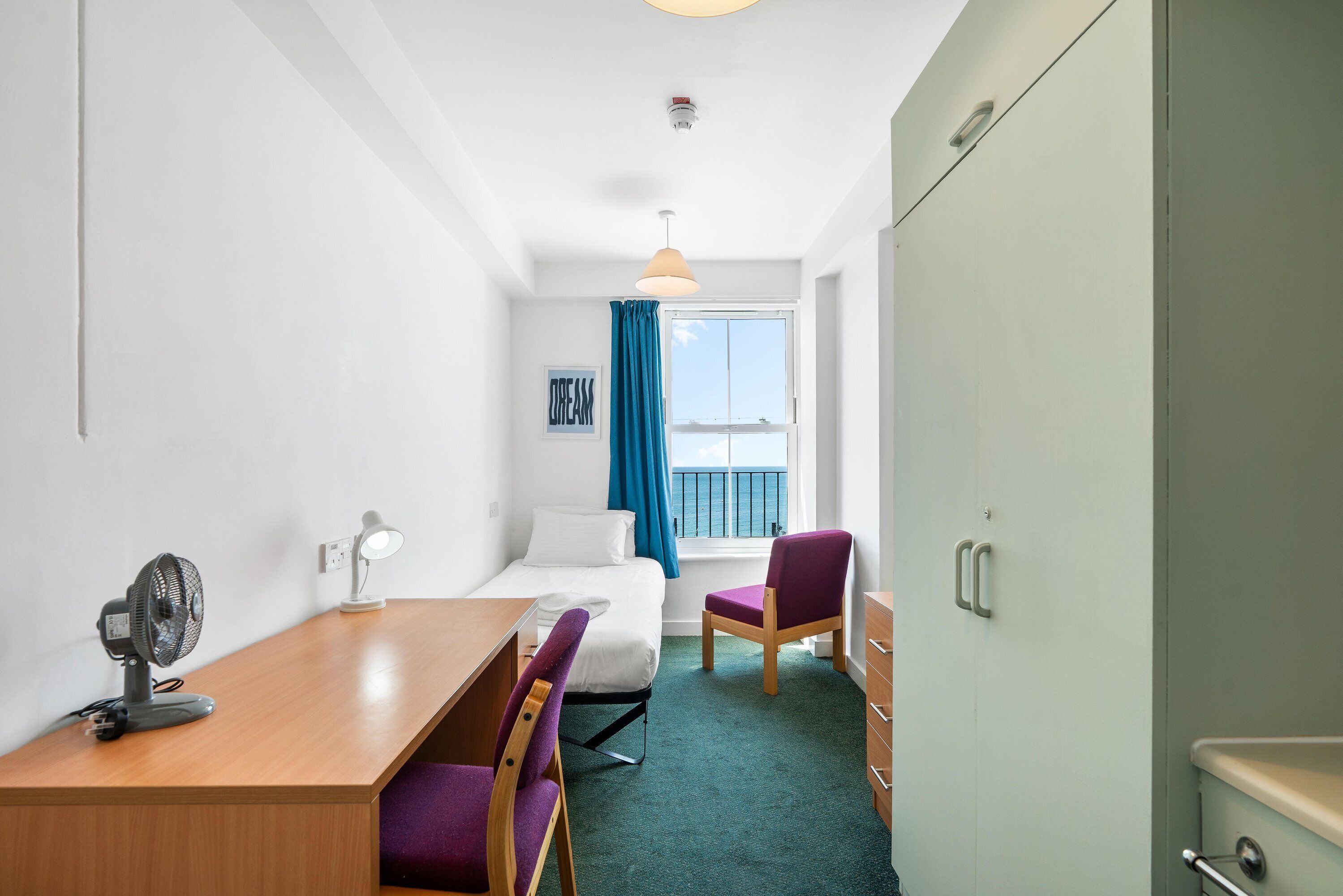 Ein Hotelzimmer mit einem Bett, einem Schreibtisch mit Ventilator, einem Sessel, einem Kleiderschrank und einem Fenster mit Blick.
