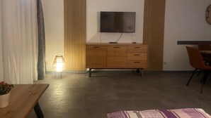 Ein Zimmer mit einem Holzkasten, einem an der Wand befestigten Fernseher, einem Schreibtisch mit Stuhl und einer Stehlampe.
