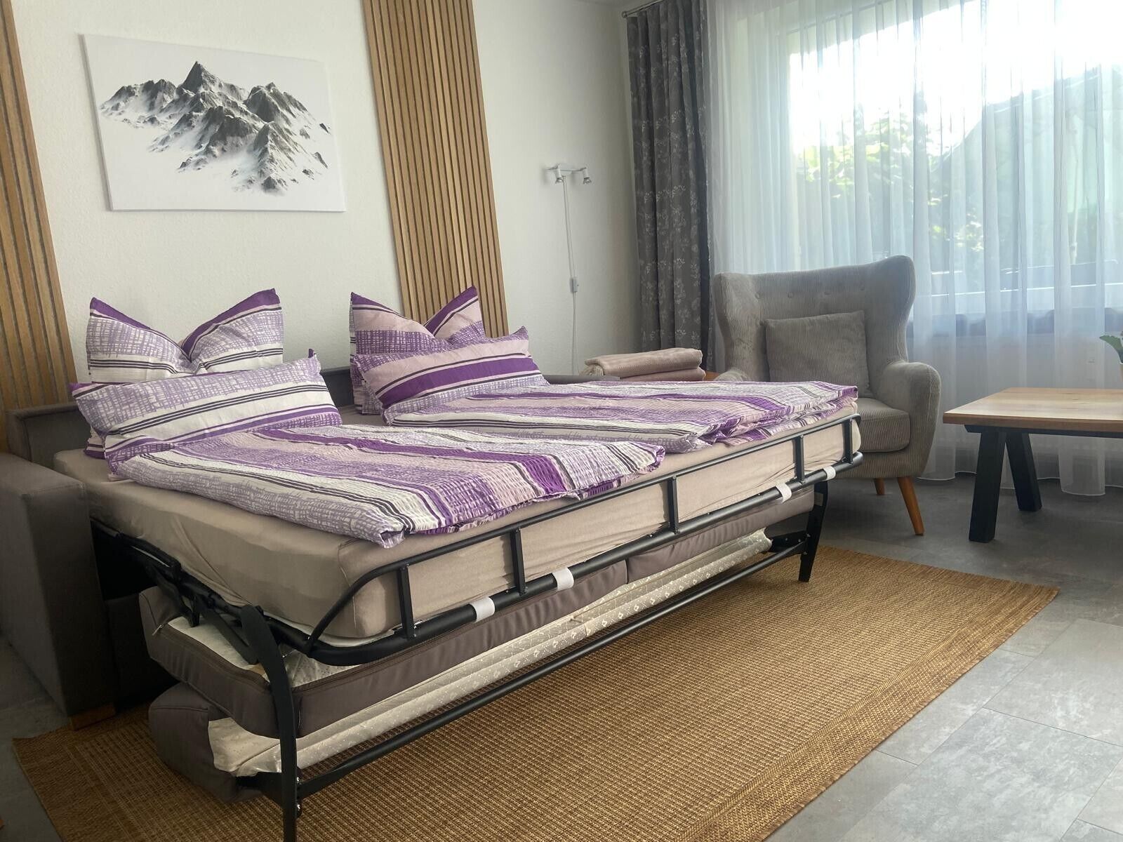 Un lit avec un cadre en métal et un tapis tissé, une chaise, une table et une fenêtre avec des rideaux.