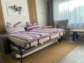 A bed with a metal frame and a woven rug, a chair, a table, and a window with curtains.