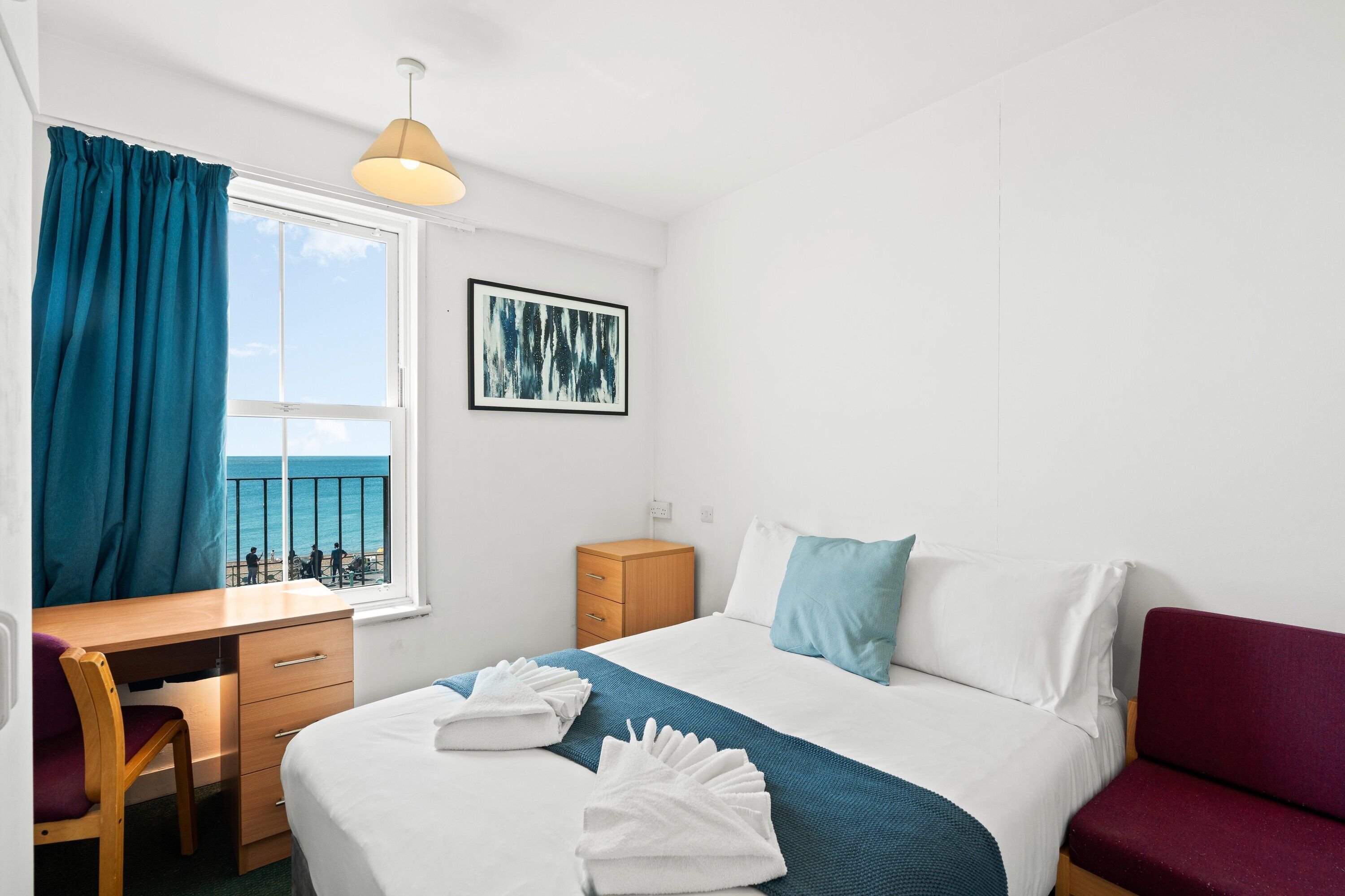 Habitación de hotel con cama, escritorio, silla, armario, ventana con cortinas y un cuadro en la pared.