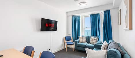 A modern living room with a flat-screen TV displaying 'NETFLIX,' a blue sofa set, a wooden dining table, and a view of the outdoors through large windows.