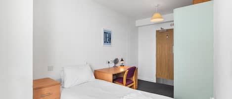 A hotel room with a single bed, a wooden nightstand, a desk with a chair, a wall-mounted fan, and a door with a wooden panel.