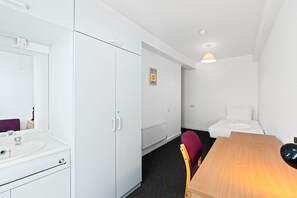 Une chambre d’hôtel avec une armoire blanche, un petit lit, une table en bois et une salle de bain équipée d’un miroir et d’un lavabo.