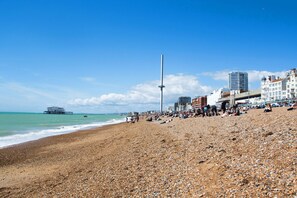 Una playa de guijarros con gente, un muelle y edificios al fondo.