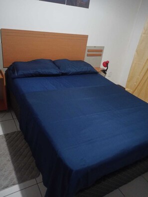 A bed with a wooden headboard, a nightstand with a red object, and a curtain partially visible. - Charming 2-bedroom house with AC in vibrant Salvador (Salvador)
