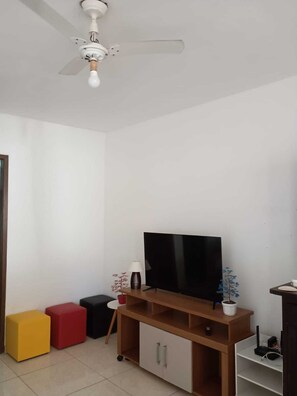 A room with a ceiling fan, a flat-screen TV on a wooden stand, a small table with a lamp, and colorful stools. - Charming 2-bedroom house with AC in vibrant Salvador (Salvador)
