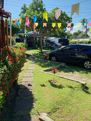 A paved pathway leading to a building with colorful flags, a parked car, and a garden with red flowers. - Charming 2-bedroom house with AC in vibrant Salvador (Salvador)