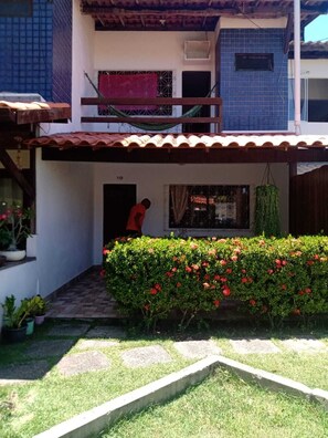 A two-story building with a balcony, a small garden with red flowers, and a person standing in the doorway. - Charming 2-bedroom house with AC in vibrant Salvador (Salvador)
