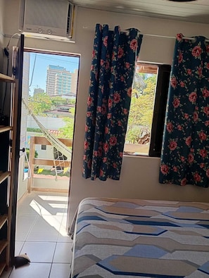 A room with a bed, a window with curtains, a radiator, and a view of buildings and greenery outside. - Charming 2-bedroom house with AC in vibrant Salvador (Salvador)