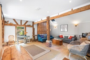 A spacious living area with exposed wooden beams, a blue sofa, a wooden coffee table, and a wicker chair. - Medleys Barn | By My Getaways (High Hurstwood)