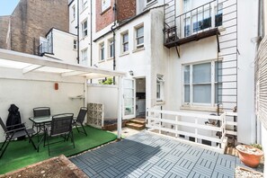 A patio with a table and chairs, a white fence, and a building with multiple windows. - Seaside Haven | By My Getaways (Brighton)