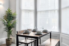 A dining table set with plates, glasses, and cutlery, positioned near a window with a view of buildings. - Seaside Haven | By My Getaways (Brighton)