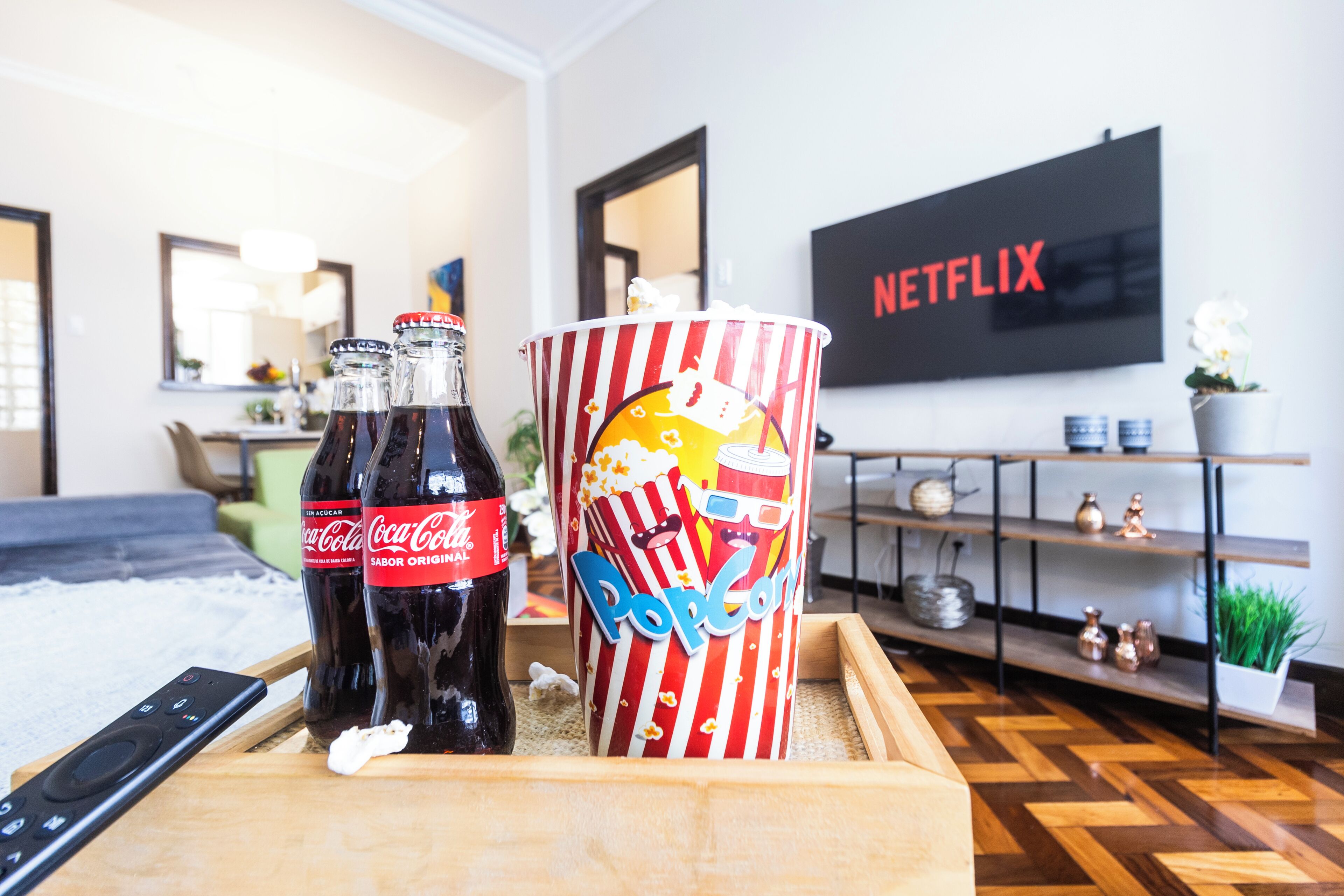 Une pièce avec une table basse en bois sur laquelle reposent deux bouteilles de Coca-Cola, un récipient à pop-corn et une télécommande. Un téléviseur diffusant Netflix est fixé au mur au fond de la pièce.