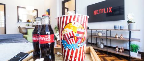 Une pièce avec une table basse en bois sur laquelle reposent deux bouteilles de Coca-Cola, un récipient à pop-corn et une télécommande. Un téléviseur diffusant Netflix est fixé au mur au fond de la pièce.