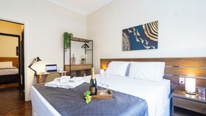 A hotel room with a bed, a desk with a laptop, a bedside table with a lamp, and a wall with a decorative piece. - 3 Quartos a 100m da Praia com AC  (Rio de Janeiro)