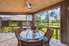 A covered outdoor area with a round glass table and wicker chairs, overlooking a tropical landscape with palm trees. - New Listing, Kanaloa, Golf Course Views, AC (Kailua-Kona)