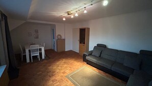 Um quarto com piso de madeira, um sofá cinza, uma mesa de jantar com cadeiras e uma luminária no teto.