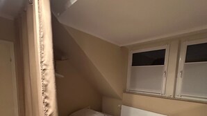 Quarto pequeno com uma cama de solteiro, teto inclinado, janela e cortina.