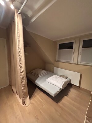 A small, single-bed room with a sloped ceiling, a window, and a curtain. - Apartment 'Familienfreundliches in Achim' with Shared Garden, Balcony and Wi-Fi (Achim)