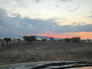 A landscape with a fence, sparse vegetation, and a distant view of a body of water and mountains during sunset.