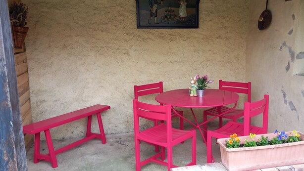 Uma pequena área externa rústica com uma mesa redonda e quatro cadeiras, um banco e um vaso com flores.