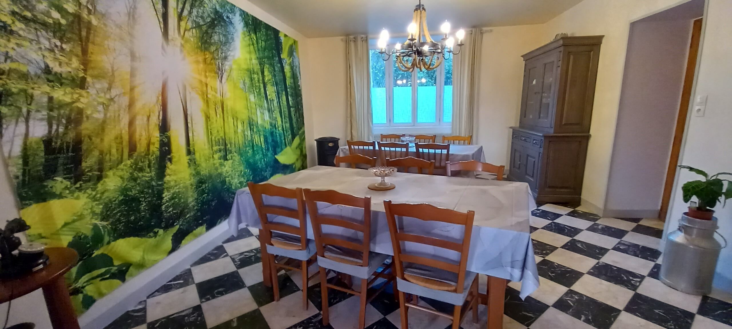 Un comedor con piso a cuadros, una mesa con sillas, una araña y un gran mural de un bosque.