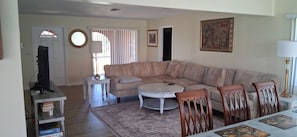Ein Wohnzimmer mit einem beigefarbenen Sofa, einem runden Couchtisch und Holzstühlen. Ein Fernseher steht auf einem Lowboard, ein kleiner Beistelltisch und ein gemustertes Teppich sind ebenfalls vorhanden.