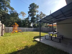 A backyard with a swing set, a picnic table, and a covered patio area. - Cozy 3BR home in the Woodlands. Perfect for families! (Tomball)