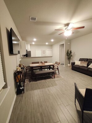 A modern living room with a dining area, a ceiling fan, recessed lighting, and a flat-screen TV mounted on the wall. - Cozy 3BR home in the Woodlands. Perfect for families! (Tomball)