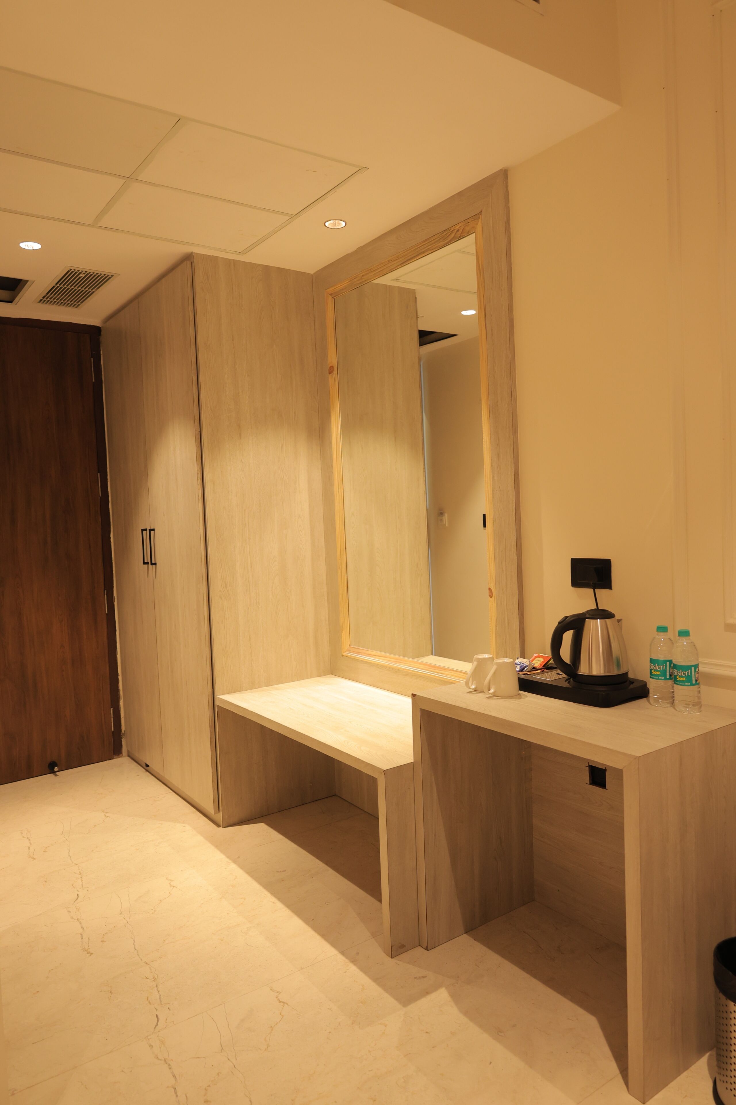 A hotel room with a wooden wardrobe, a desk with a mirror, a coffee maker, and two bottles of water.