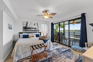 Ein Schlafzimmer mit Bett, Nachttisch, Schreibtisch, Stuhl, einem Balkon mit Blick auf das Wasser und einem Deckenventilator.