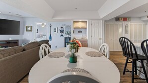Un coin repas avec une table ronde, des chaises et un vase rempli de fleurs. On aperçoit, au fond, une cuisine avec des placards et un évier.